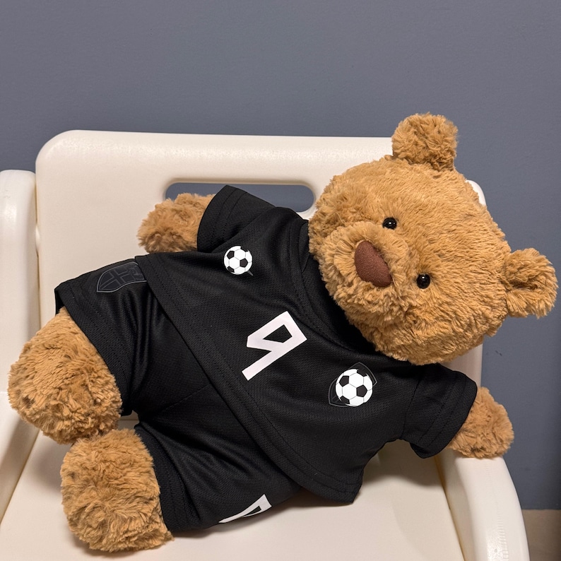May include: A light brown plush teddy bear wearing a black athletic jersey and matching shorts. The jersey features a white number 9, a small soccer ball graphic, and a shield emblem. The fabric has a textured, mesh-like appearance.