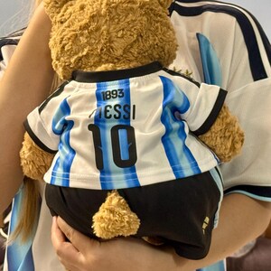 May include: A brown plush teddy bear wearing a white and light blue striped soccer jersey with the text 1893, MESSI, and the number 10 printed on the back, paired with black shorts that have a small gold logo on the lower left side.