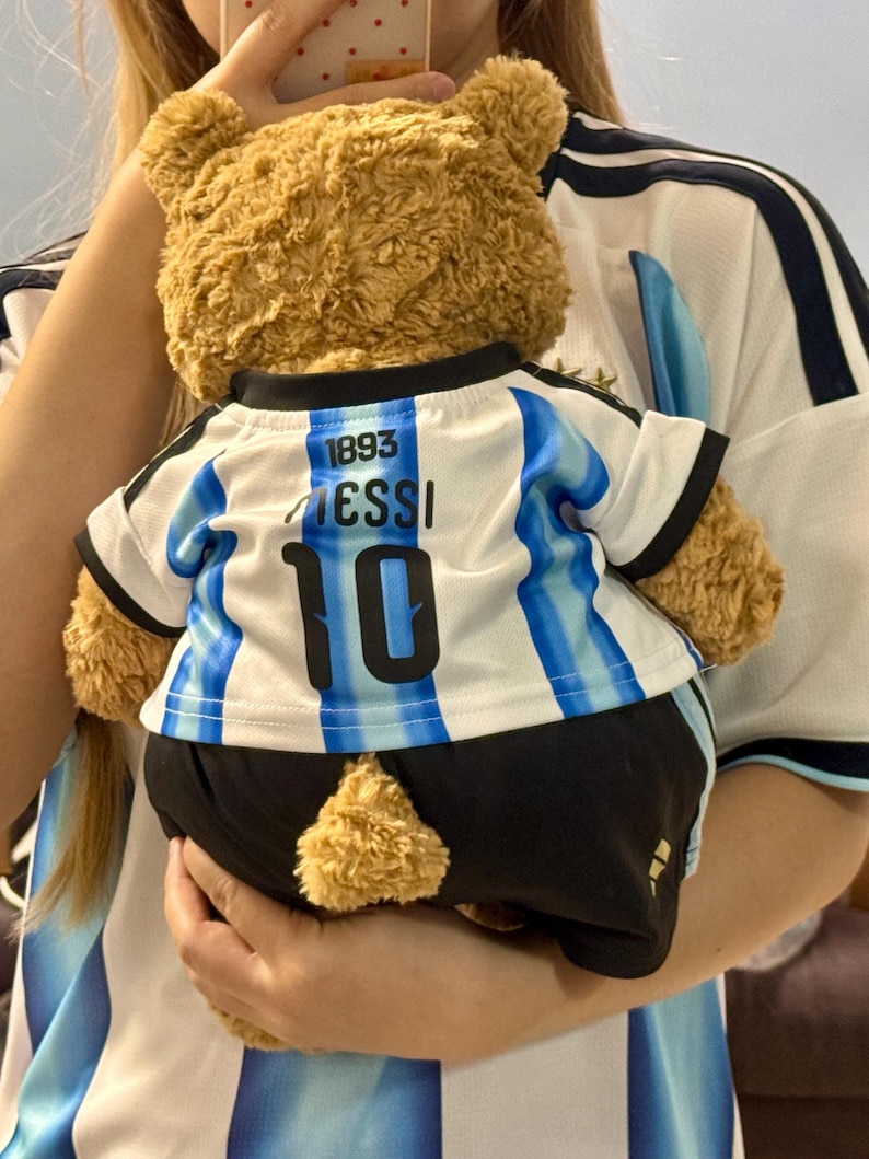 May include: A brown plush teddy bear wearing a white and light blue striped soccer jersey with the text 1893, MESSI, and the number 10 printed on the back, paired with black shorts that have a small gold logo on the lower left side.