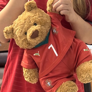 May include: A brown plush teddy bear wearing a red soccer jersey with a dark green collar, the number 7 in white, and the Portugal national team crest. The jersey features small logos and the word cool printed on the chest and leg area.