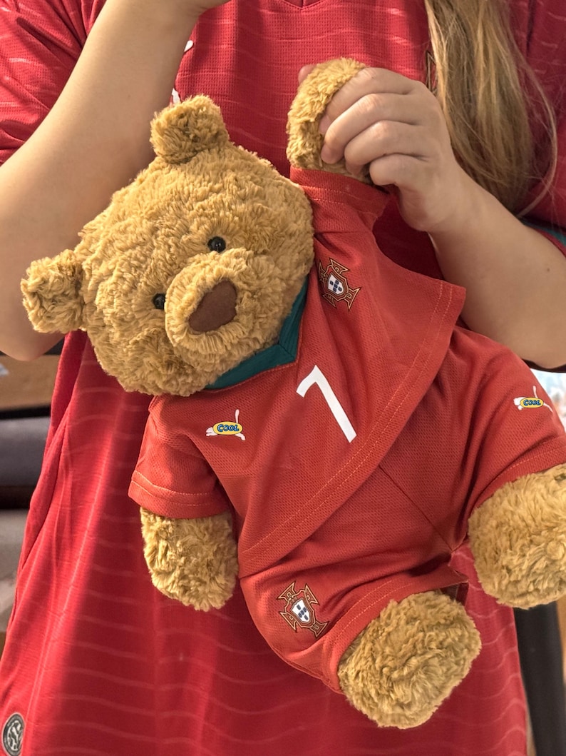 May include: A brown plush teddy bear wearing a red soccer jersey with a dark green collar, the number 7 in white, and the Portugal national team crest. The jersey features small logos and the word cool printed on the chest and leg area.