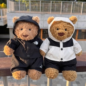 May include: Two brown plush teddy bears wearing hooded jackets and trousers. The bear on the left wears a solid black outfit with a white logo on the chest. The bear on the right wears a white and black colour-blocked jacket with black trousers.