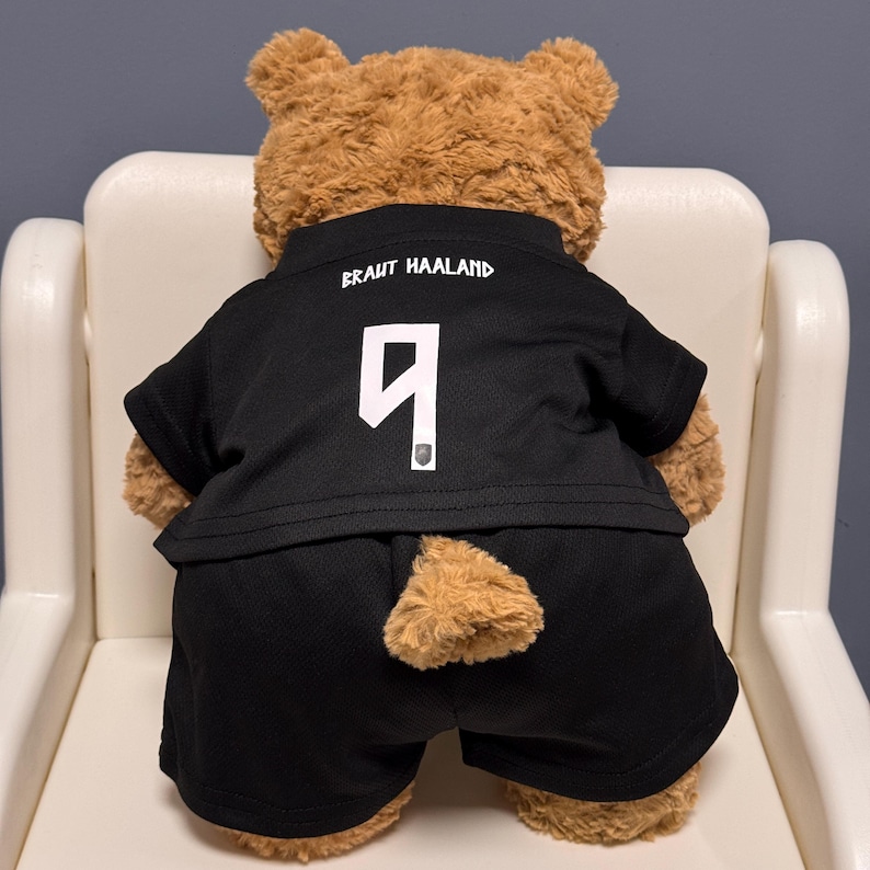 May include: A brown plush teddy bear wearing a black athletic jersey and matching black shorts. The jersey features the white text BRAUT HAALAND and the number 9 printed on the back. The bear is viewed from the rear, showing its fuzzy tail.