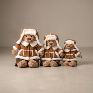 May include: Three teddy bears dressed in brown aviator jackets with white faux fur trim and matching hats. The bears are of different sizes, standing together. The bears have brown fur and are wearing brown trousers.
