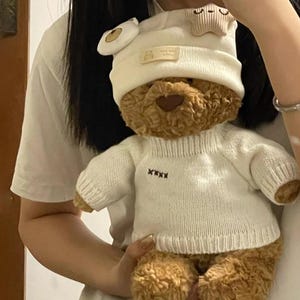 Op de afbeelding: Een bruine teddybeer met een witte trui en bijpassende hoed. De hoed heeft een ster en een berengezicht. De trui heeft een gestikt ontwerp op de voorkant. De beer is een knuffel.