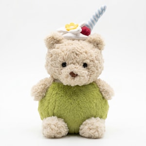 Puede incluir: Un oso de peluche con pelaje marrón claro, vistiendo un atuendo verde. El oso tiene un sombrero blanco con una flor amarilla y una baya roja, y un cuerno a rayas azules y blancas. El oso está sentado sobre un fondo blanco.