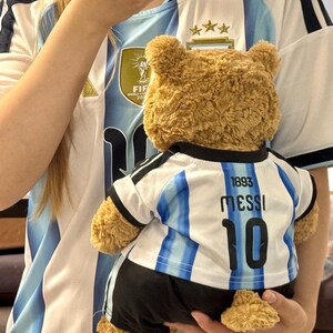 May include: A brown plush teddy bear wearing a white and light blue striped soccer jersey with black trim. The back of the jersey features the text 1893, the name MESSI, and the number 10 in black lettering, paired with black athletic shorts.