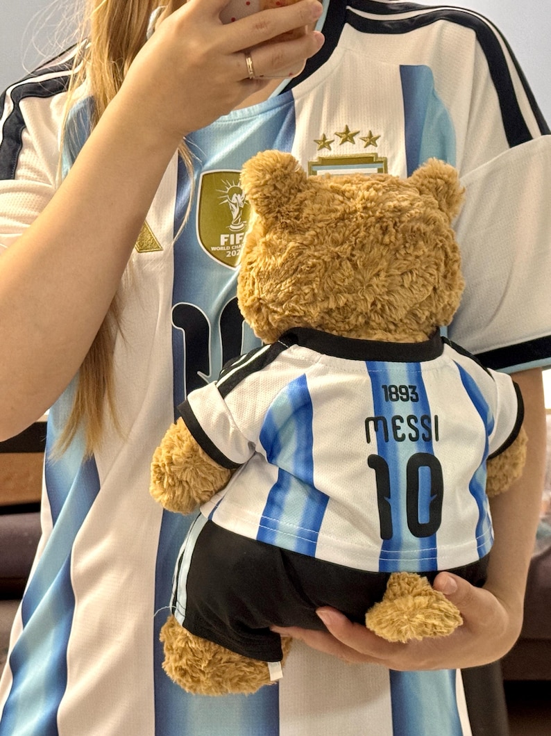 May include: A brown plush teddy bear wearing a white and light blue striped soccer jersey with black trim. The back of the jersey features the text 1893, the name MESSI, and the number 10 in black lettering, paired with black athletic shorts.