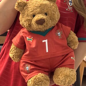 May include: A brown plush teddy bear wearing a red soccer jersey and matching shorts. The jersey features a white number 1, a green V-neck collar, a small 'COOL' patch, and the Portugal national team crest. The shorts also display the team crest.