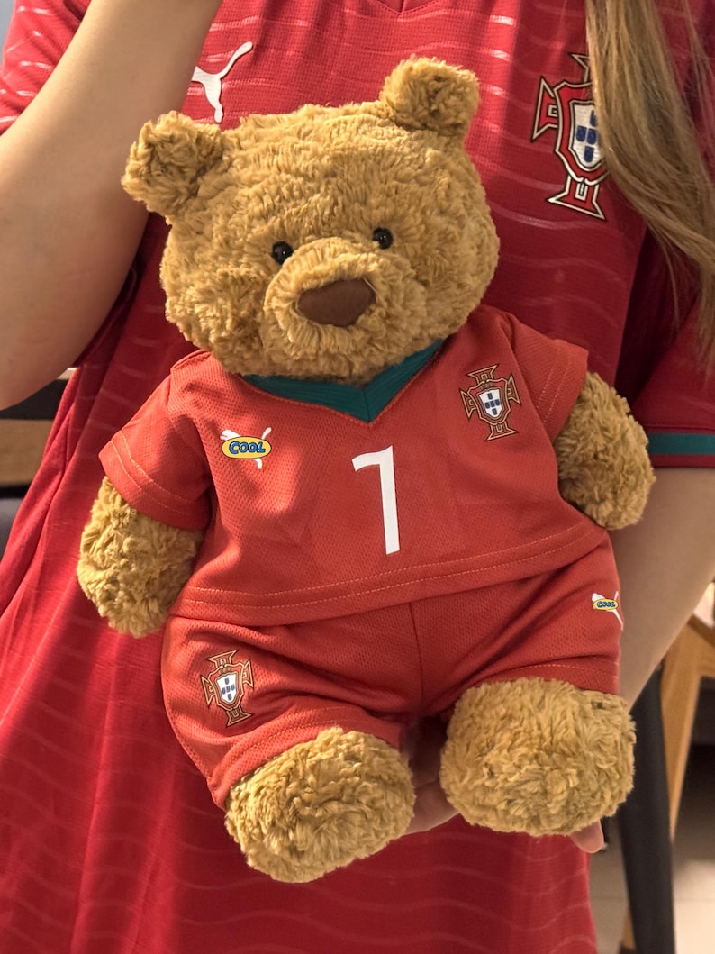 May include: A brown plush teddy bear wearing a red soccer jersey and matching shorts. The jersey features a white number 1, a green V-neck collar, a small 'COOL' patch, and the Portugal national team crest. The shorts also display the team crest.