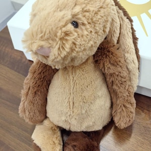 May include: A plush bunny toy in shades of brown and tan. The bunny has long, floppy ears, a small pink nose, and dark eyes. The soft toy is sitting on a wooden surface.