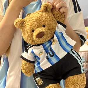 May include: A light brown plush teddy bear wearing a white and light blue striped soccer jersey with black shorts. The jersey features the AFA logo on the chest, the word Cool on the right side, and the AFA logo on the right leg of the shorts.