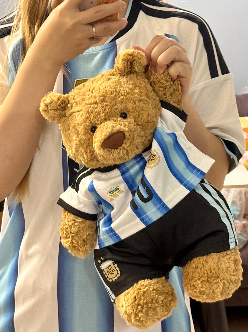 May include: A light brown plush teddy bear wearing a white and light blue striped soccer jersey with black shorts. The jersey features the AFA logo on the chest, the word Cool on the right side, and the AFA logo on the right leg of the shorts.