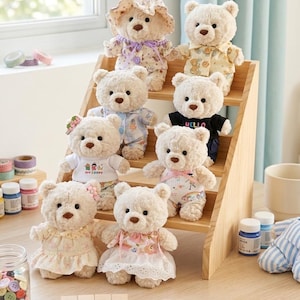 May include: A collection of plush teddy bears in various outfits, including dresses, pajamas, and a shirt that says "HELLO". The bears are arranged on a wooden display shelf. The word "HAND MADE" is spelled out with wooden blocks.