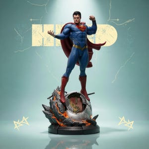 Puede incluir: Una figura de Superman en una pose dinámica, vestida con un traje azul con capa y botas rojas. La figura se alza sobre una base detallada con un diseño metálico y ardiente. La palabra "HERO" es visible en el fondo.
