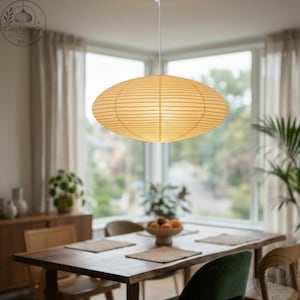 May include: An oval-shaped, paper lantern pendant light fixture hangs above a wooden dining table. The light is a warm, soft yellow, and the fixture has horizontal lines. The room has large windows and neutral-colored curtains.