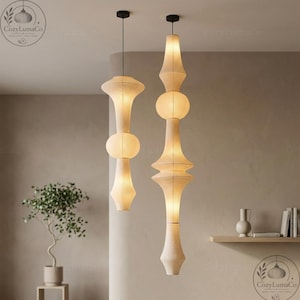 May include: Two beige pendant lights with black fittings hang from the ceiling. The lights have a layered design with a cylindrical base, a spherical middle, and a flared top. The lights are in a neutral-toned room.