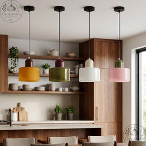 Può includere: Quattro lampade a sospensione con paralumi cilindrici gialli, verde oliva, bianchi e rosa. Ogni lampada ha un cavo nero e un supporto a soffitto nero. Le lampade sono appese in una cucina con mobili e mensole in legno.