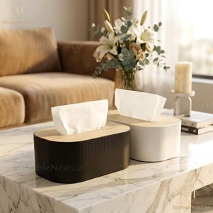 Puede incluir: Dos cajas de pañuelos, una negra y otra blanca, con una tapa de madera clara. El soporte negro es rectangular con ranuras verticales, y el blanco es ovalado. Ambos tienen pañuelos blancos que emergen de la parte superior.