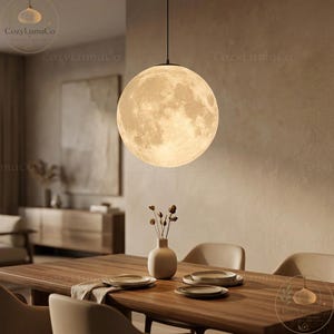 May include: A round, moon-shaped pendant light hangs above a wooden dining table. The light fixture has a detailed surface resembling the moon's craters. A vase with dried flowers and place settings are on the table. The room has a neutral color palette.