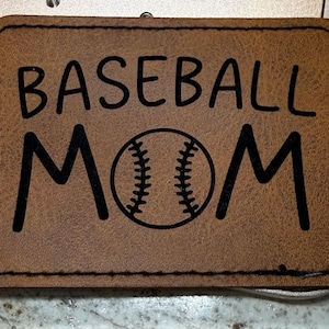 Könnte beinhalten: Ein rechteckiger, brauner Lederaufnäher mit den Worten "BASEBALL MOM" in fetten, schwarzen Buchstaben. In der Mitte befindet sich eine Baseball-Grafik. Der Aufnäher hat einen genähten Rand.