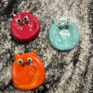 May include: Three brightly coloured cat-shaped ornaments. The cats are red, turquoise, and orange, each with large eyes, small ears, and black whiskers. They appear handmade and are on a speckled grey surface.