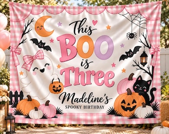 May include: A rectangular fabric backdrop featuring a pink gingham border and a white center with the text This BOO is Three Madeline's SPOOKY BIRTHDAY. The design includes a ghost with a bow, pumpkins, a black cat, bats, and a crescent moon.