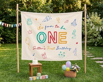 以下が含まれることがあります： クリーム色の布製背景幕。カラフルな文字で「it's fun to be ONE」「Sutton's First Birthday」と書かれ、ケーキ、パーティーハット、風船、ギフト、花火のイラストが描かれており、2本の木の棒の間に吊り下げられています。