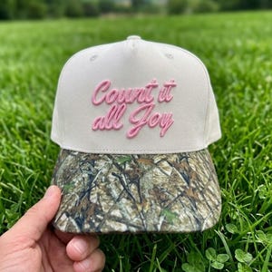 May include: A baseball cap with a cream-colored front panel featuring the embroidered pink text "Count it all Joy" in a cursive font. The brim of the cap displays a detailed woodland camouflage pattern in shades of green, brown, and tan.