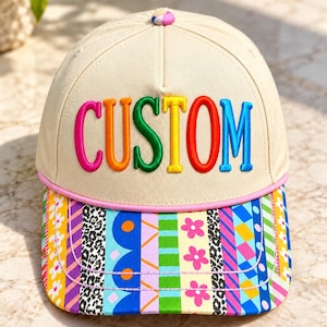 May include: A beige baseball cap featuring the word CUSTOM embroidered in multi-colored letters across the front. The brim displays a vibrant pattern of vertical stripes, including floral, leopard print, and geometric designs, with a pink trim.
