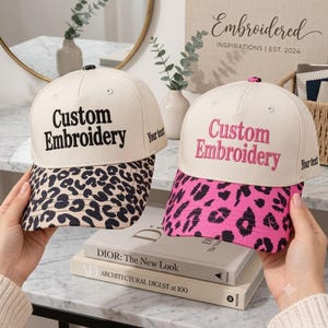 May include: Two baseball caps with off-white crowns and leopard print brims. The left cap features black embroidered text reading Custom Embroidery and Your text. The right cap features pink embroidered text reading Custom Embroidery and Your text.