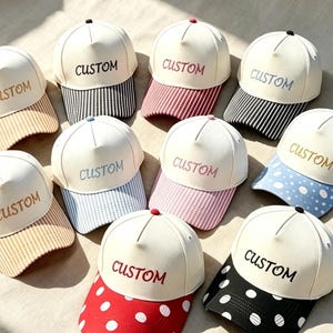 May include: A collection of ten baseball caps with off-white crowns and the word CUSTOM embroidered on the front. The brims feature various patterns including black and white stripes, red and white stripes, blue and white polka dots, and red with white dots.