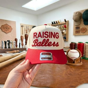 May include: A baseball cap with a red brim and a cream-colored crown. The text "RAISING Ballers" is embroidered in red. The cap has a classic fit and profile.
