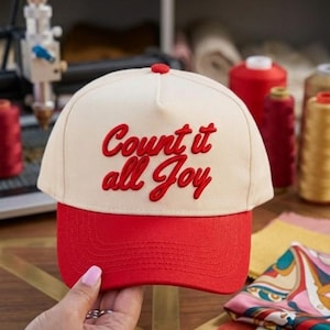 May include: A cream-colored baseball cap with a red brim and red embroidered text on the front that reads Count it all Joy. The cap features a structured crown, a top button, and stitched detailing on the brim, showcasing a two-tone design.