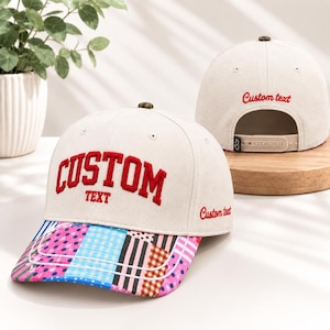 May include: An off-white baseball cap featuring red embroidered text that reads CUSTOM TEXT on the front and side. The brim displays a colorful patchwork pattern of polka dots, gingham, and stripes. A second cap is visible in the background.
