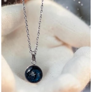 May include: Silver-toned necklace with a circular pendant. The pendant features a dark blue background with a white snowflake design. The necklace chain is delicate and hangs from the pendant.