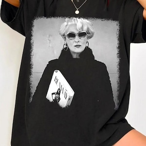 May include: Black t-shirt featuring a black and white image of a woman wearing sunglasses and holding a book titled "Runway". The image has a white border. The t-shirt is a casual, oversized style.
