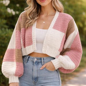 May include: A cropped, patchwork cardigan in shades of pink, white, and brown. The sweater has long, puffy sleeves and a V-neck. The model is wearing light blue jeans and a white top. The cardigan is a cozy, stylish piece.