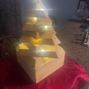 May include: A wooden Christmas tree with a geometric design. The tree is constructed from light-colored wood and features built-in warm white LED lights. It sits on a red tablecloth, creating a festive holiday decoration.