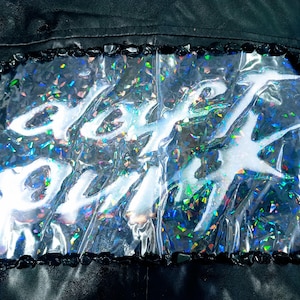 May include: Black leather jacket detail with a clear, iridescent panel featuring the white text "DAFT PUNK". The panel is outlined with black stones, creating a striking contrast against the jacket's dark background.