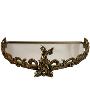 May include: An antique-style shelf with a mirrored surface and ornate, swirling bronze-colored metalwork. A small fairy statue is centered on the shelf. The shelf is designed to be mounted on a wall.