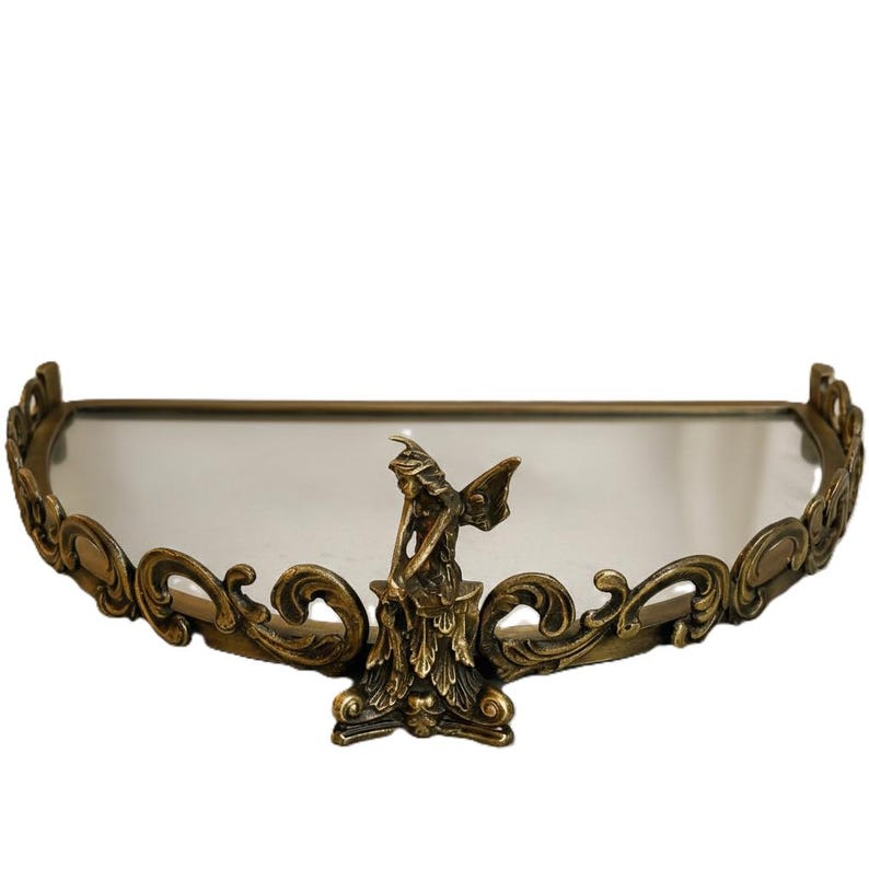 May include: An antique-style shelf with a mirrored surface and ornate, swirling bronze-colored metalwork. A small fairy statue is centered on the shelf. The shelf is designed to be mounted on a wall.