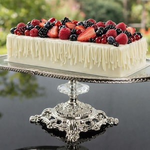 Puede incluir: Un pastel rectangular blanco con un glaseado blanco, cubierto con bayas frescas, incluyendo fresas, frambuesas, arándanos y moras. El pastel está sobre un soporte plateado y ornamentado con un acento de cristal claro.