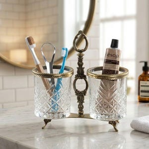 May include: A decorative toothbrush and toothpaste holder with a vintage design. The holder has two clear, patterned glass cups with gold-colored rims, connected by an ornate metal stand. The holder contains toothbrushes, a tongue scraper, and a tube of toothpaste.