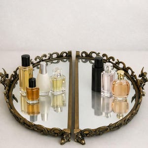 May include: An oval, antique-style mirrored tray with a decorative bronze-colored frame. The tray holds several perfume bottles of various shapes and sizes, with black, silver, and gold caps. The bottles reflect in the mirror.