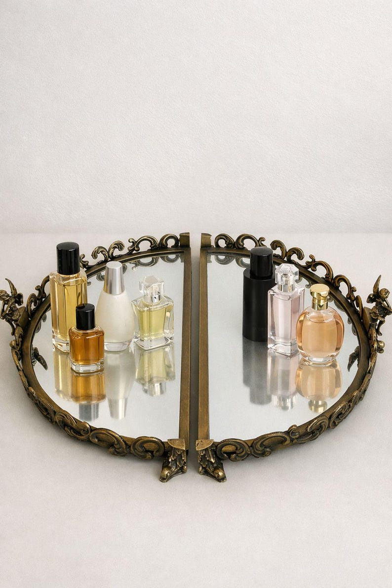May include: An oval, antique-style mirrored tray with a decorative bronze-colored frame. The tray holds several perfume bottles of various shapes and sizes, with black, silver, and gold caps. The bottles reflect in the mirror.