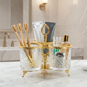 May include: A decorative bathroom organizer with three sections. The organizer is made of clear, cut glass with gold-colored metal accents. It holds toothbrushes, toothpaste, and other toiletries. The organizer sits on a marble countertop.