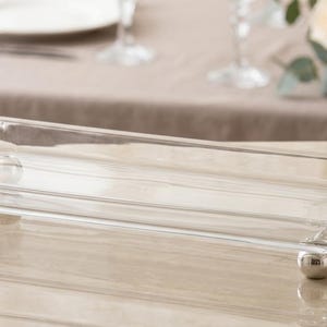 Könnte beinhalten: Ein rechteckiges Serviertablett aus klarem Glas mit verzierten silbernen Griffen und Kugelfüßen. Das Tablett steht auf einer reflektierenden Oberfläche, mit verschwommenem Hintergrund.