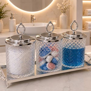 Puede incluir: Tres frascos de vidrio transparente con textura de diamante y tapas plateadas con asas decorativas. Los frascos se asientan sobre una bandeja rectangular plateada con efecto espejo y asas ornamentadas, conteniendo polvo blanco, cápsulas de detergente y líquido azul.