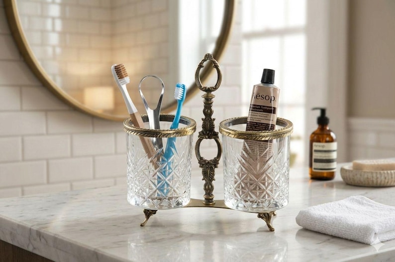 May include: A decorative toothbrush and toothpaste holder with a vintage design. The holder has two clear glass cups with a diamond pattern, trimmed with gold. The holder contains toothbrushes, a tongue scraper, and a tube of toothpaste.